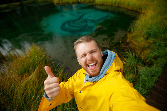 Happy travel selfie of man enjoying nature by turquoise Blue geyser lake in bright yellow raincoat Altay Russia