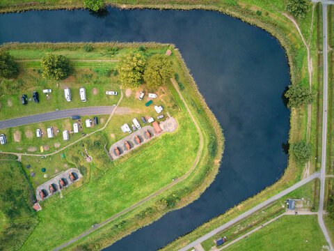 Aerial view of a tranquil campsite nestled by a winding river bend, with rows of white caravans contrasting against the vibrant green grass, Gentofte, Denmark.