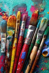 Fototapeta premium Colorful paintbrushes, various sizes and colors, stained with vibrant pigments, resting on a teal surface covered in paint splatters