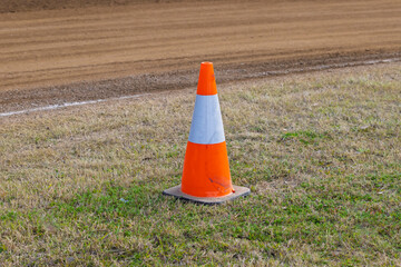 Photograph of an industrial orange safety cone located on the infield corner of a dirt speedway race track on a sunny day. 