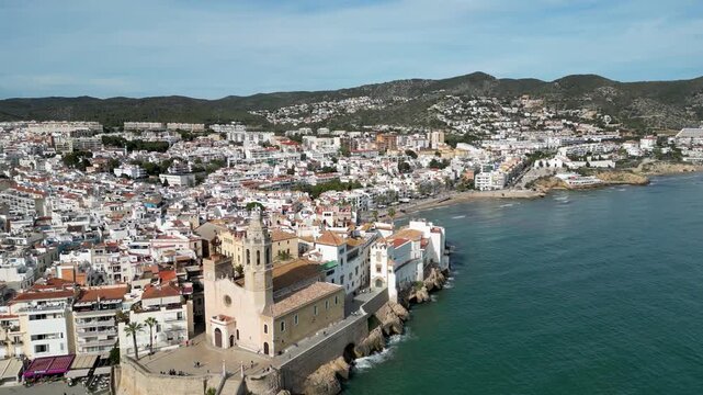 Aerial view of the Church of Sant Bartomeu and Santa Tecla's golden facade against the turquoise waters, creating a stunning contrast, Sitges, Barcelona, Spain.