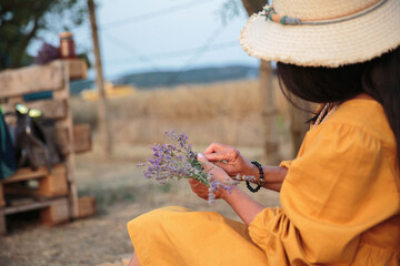 Mature woman in straw hat and yellow dress creating a lavender bouquet outdoors with scenic...