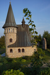 Fototapeta premium a stone church with a tall steeple and a clock on it. The church has a sloped roof and is situated on a grassy hill.