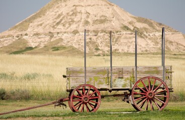 Vintage wooden wagon with weathered red wheels and faded green paint, set against a backdrop of golden prairie grass and a striking butte in the American Great Plains. Evokes the spirit of the Old Wes