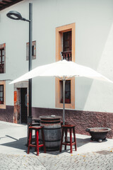 Cozy outdoor seating with a rustic wooden barrel table, three red stools, and a white umbrella in front of a traditional Spanish building with textured walls and shuttered windows, sunny day