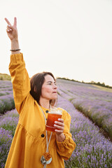 Confident woman in yellow dress raising hand with peace sign while holding honey jar in blooming...