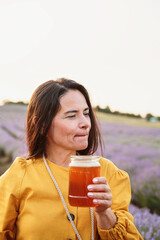 Pensive woman with glass jar of honey standing in blooming purple lavender field during sunset