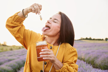 Cheerful woman holding lavender sprig with honey above mouth in countryside during golden hour