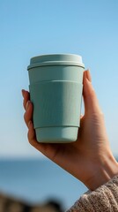 Reusable coffee cup in dill green silicone sleeve held by manicured hand against blue sky  