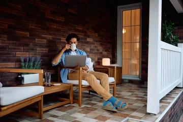  Relaxed man using computer while drinking coffee on porch. © Drazen