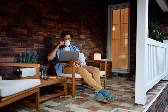 Relaxed man using computer while drinking coffee on porch.