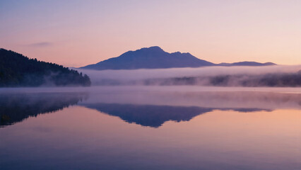 Pastel reflection of mountain silhouette on mist-covered lake under pale pink sky
