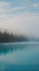 Mist hovering above still forest lake, soft aqua color palette and light cloud cover.