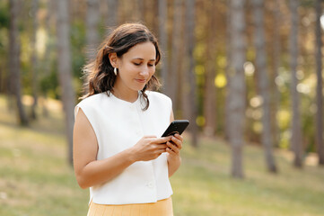 A young woman in a serene forest setting, engrossed in her smartphone, exemplifying modern lifestyle.