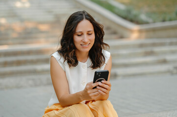 A woman is sitting on steps outdoors, joyfully smiling at her smartphone while enjoying a peaceful moment in nature