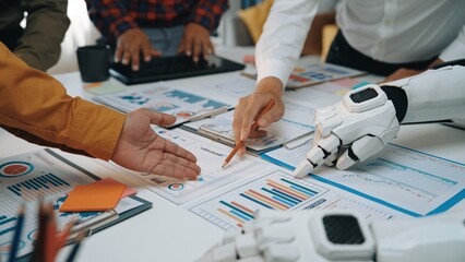 Professionals engage in a collaborative effort, using both human and robotic hands to analyze detailed business charts in a modern office environment, emphasizing innovation.Noogenesis