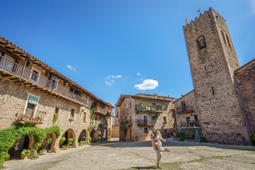 Romantic medieval main square Plaça Major de Santa Pau, Spain, with historic stone arcades, rustic balconies, and iconic watchtower under blue sky. Ideal for couples’ travel, heritage or honeymoon