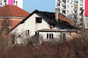 A white house with extensive fire damage and a partially collapsed roof sits abandoned in an urban...