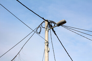 A concrete utility pole supports multiple tangled power and communication lines branching in various directions, topped with a modern streetlamp under a blue sky