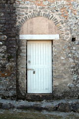 A weathered stone wall encloses a sturdy white wooden door secured by a padlock, topped with an arched stone lintel, evoking history and rustic architecture