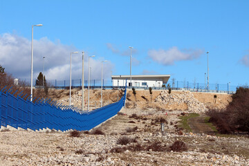 A high blue security fence topped with spikes leads to a modern building on a hill, surrounded by...