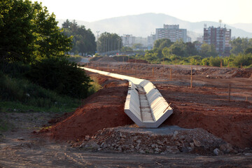 A newly assembled line of large precast concrete drainage channels stretches through red earth, forming part of a new urban infrastructure project near city buildings and mountains