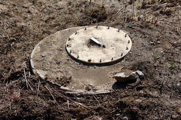 A heavy concrete manhole cover with metallic bolts and a handle is installed on rough terrain, marking access to underground utilities at a developing construction site