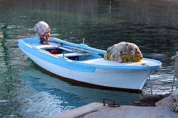A blue and white fishing boat, equipped with coiled nets and ropes, floats quietly on tranquil greenish harbor water, ready for the daily tasks of traditional coastal fishing