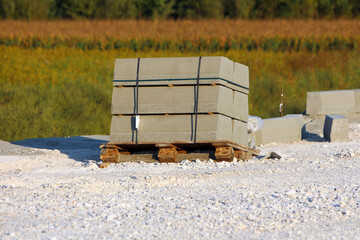 A bundle of rectangular precast concrete curbs, bound with black straps, sits atop a wooden pallet surrounded by gravel at a roadwork construction site
