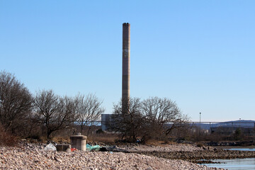 A towering smokestack rises above industrial buildings at the edge of a refinery, near a rocky shoreline and bare trees, set against a pale blue sky and distant hills