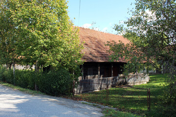 A rustic wooden house with red tile roofing sits shaded by trees and surrounded by a picket fence, blending traditional rural architecture with verdant summer growth