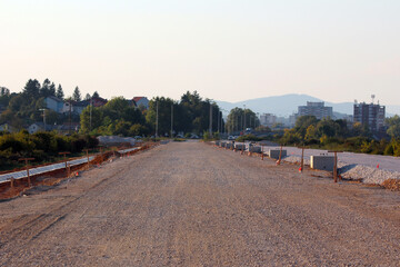 A long stretch of unpaved road foundation with placed and yet-to-be-installed curbstones lines both sides, showing active civil engineering and preparation for asphalt paving