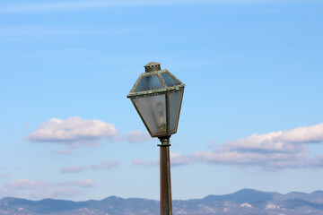 An old, weathered metal street lamp rises into a clear blue sky, with soft clouds and a mountainous landscape forming a picturesque and tranquil outdoor backdrop