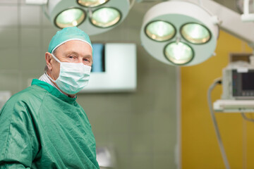 Senior male surgeon wearing green scrubs and mask standing under surgical lights in operating room