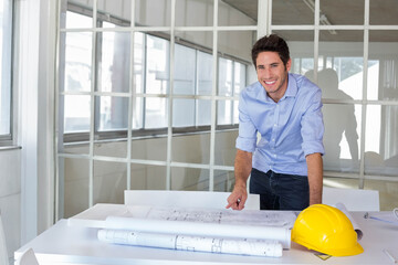 Male architect in twenties reviewing blueprints at office desk with yellow hard hat, copy space