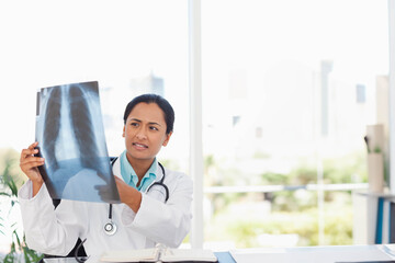 Indian woman doctor examining chest X-ray film in white lab coat at clinic desk with stethoscope