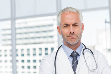 Senior male doctor standing at clinic wearing white lab coat and stethoscope by windows, copy space