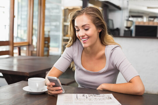 Woman sitting at wooden table inside cafe wearing gray top smiling at smartphone beside coffee cup
