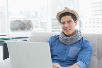 Man wearing straw hat, gray scarf sitting on sofa by windows using laptop, copy space