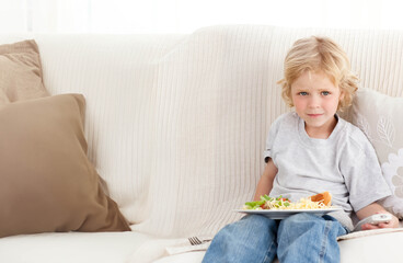 Boy holding remote control and eating salad pasta bread on sofa in living room, copy space
