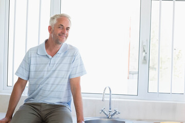 Man sitting on tiled kitchen countertop next to stainless steel sink faucet by window, copy space