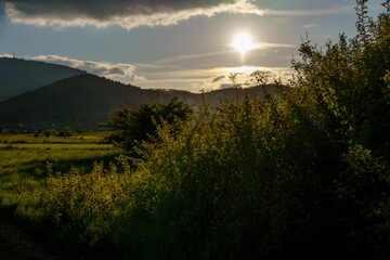 sunset behind mountains with blue sky, white clouds, and shadows on bushes in foreground