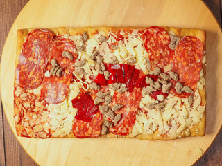 Meat feast pizza with different kind of salami and ham in rectangular shape on wooden background. Popular Italian dish with dough base, tomato sauce, cheese and other ingredients and spices.