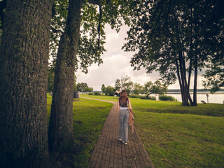 Fototapeta premium A woman walks down a path in a park. The path is lined with trees and the sky is cloudy. there is lake on the right and cloudy sky. Going on a blind date in a public place concept.