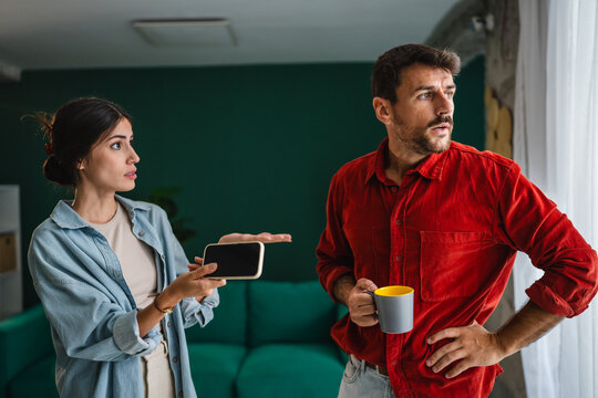 Young couple arguing at home holding smart phone and coffee cup