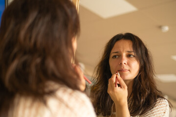 Woman applying lipstick while looking into a mirror in a bright indoor setting during the day