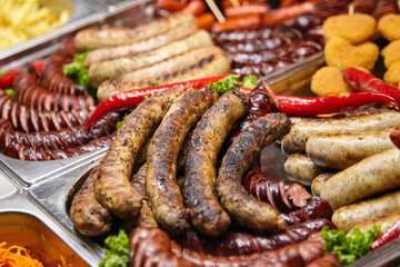 Grilled sausages of various types arranged on a metal tray with fresh herbs and red chili peppers, showcasing a vibrant display of culinary delights and textures