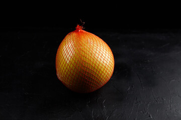 Pomelo fruit wrapped in a bright orange netting sits on a dark textured surface, showcasing its...