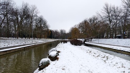 The partially ice-covered canal runs through the park among willows and poplars. The banks are concreted. Nearby boulders, lampposts and buildings. Snow covers the ground. Overcast winter weather