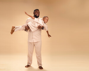 In this wide studio shot, an African father lifts his son like a soaring plane. Their matching...